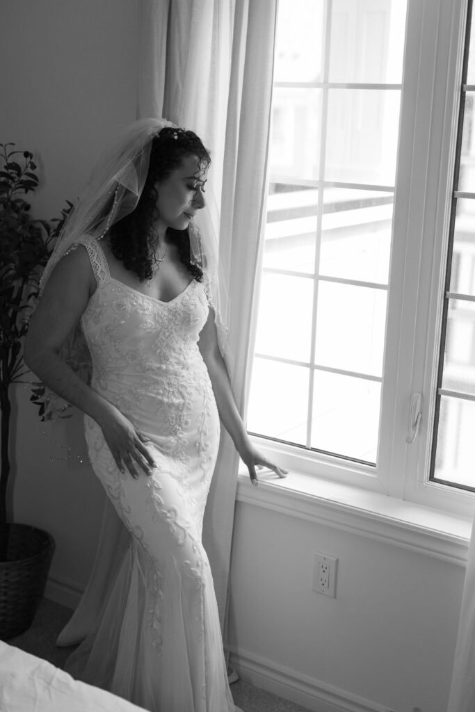 A bride in a lace wedding dress and veil stands gazing out a window in a softly lit room, her hand resting on the windowsill. Black and white photo.