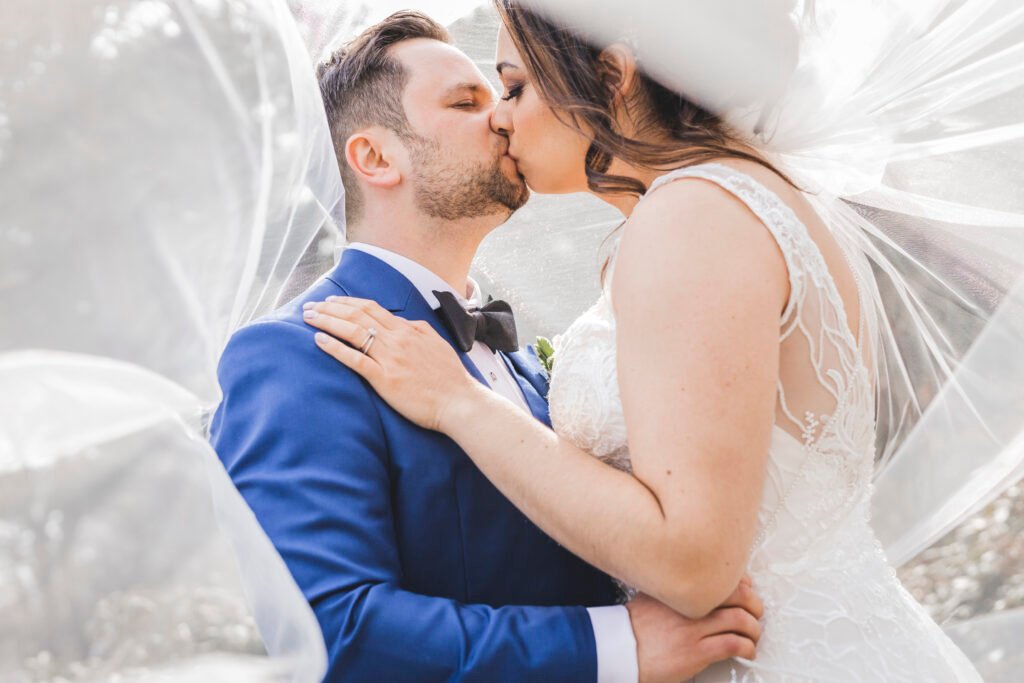A bride and groom share a kiss under a flowing veil. The groom wears a blue suit with a bow tie, and the bride wears a lace wedding dress, gently holding his shoulder. The soft veil frames their intimate moment.